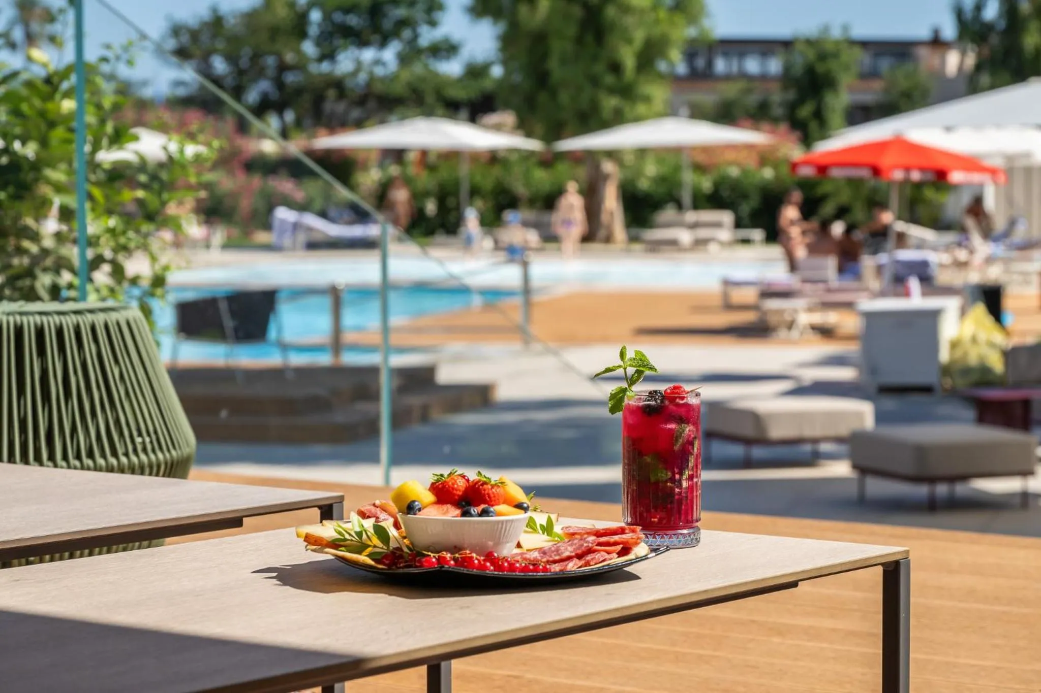 Une table d'extérieur présente une assiette de fruits et une boisson rafraîchissante près d'une piscine ensoleillée.