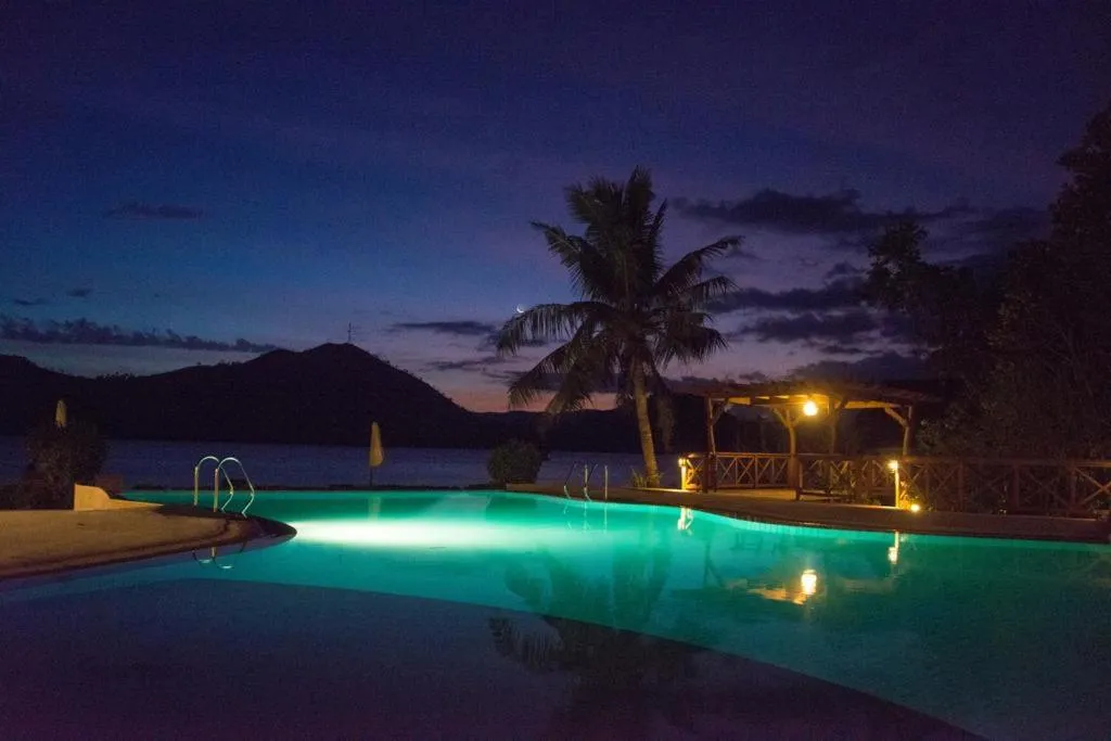 Une piscine illuminée au crépuscule, avec des palmiers et une vue sur la mer.