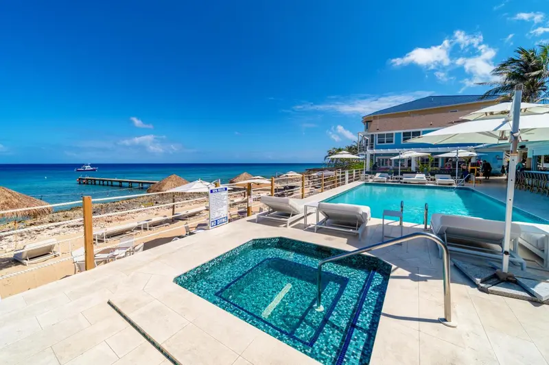 Une piscine turquoise avec des transats et un jacuzzi au premier plan, bordée d'une plage de sable et d'une mer bleue.