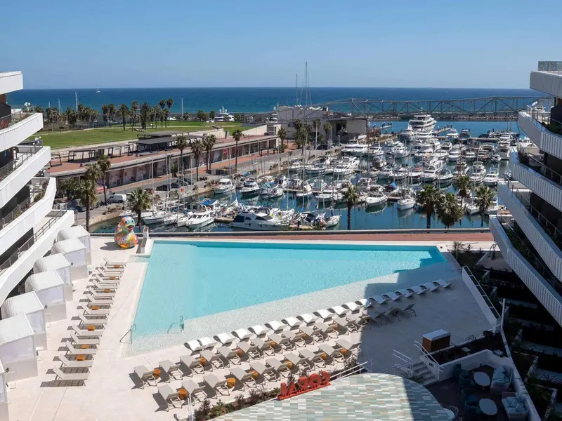 Vue aérienne d'une piscine d'hôtel avec des transats et un port de plaisance en arrière-plan.