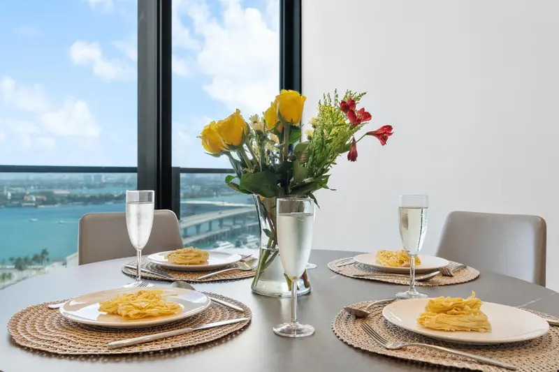 Une table est dressée avec des assiettes, des couverts et des boissons, devant une grande fenêtre avec vue sur la mer.