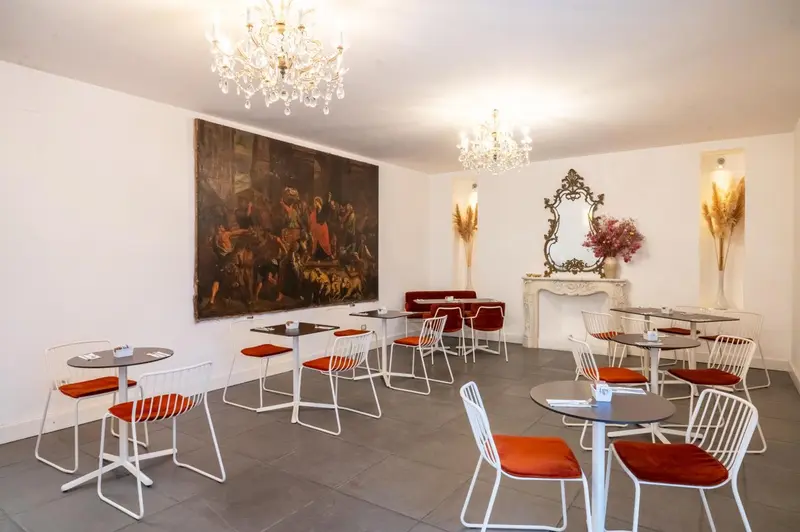 Une salle lumineuse avec des tables et des chaises, décorée de lustres et d'un grand tableau.