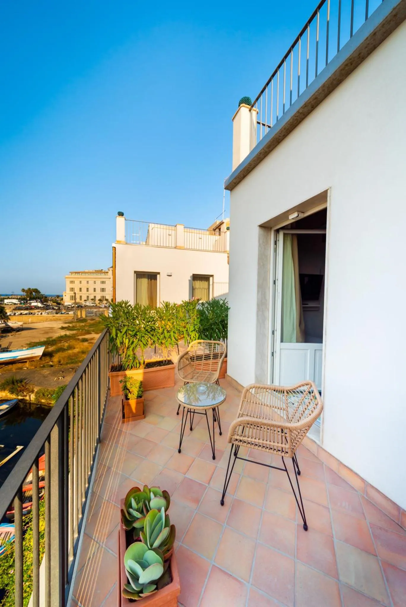Terrasse avec mobilier en rotin et plantes en pot offrant une vue sur la mer et les bâtiments environnants.
