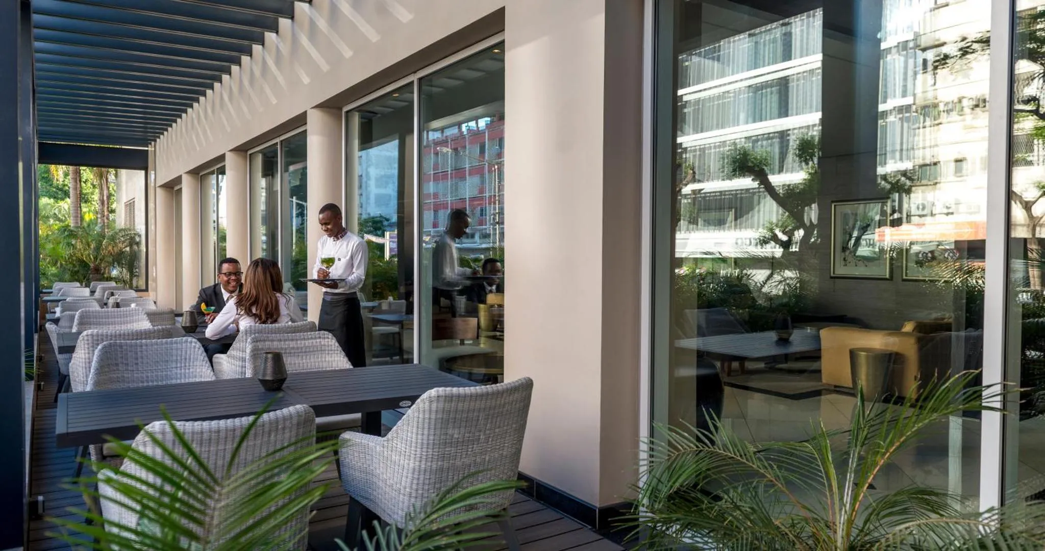 Une terrasse ombragée avec des tables et des chaises accueillant des clients servis par du personnel.