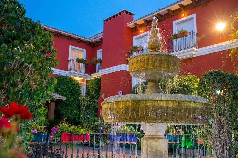Une fontaine illuminée orne une cour intérieure avec une végétation luxuriante et un bâtiment rouge.