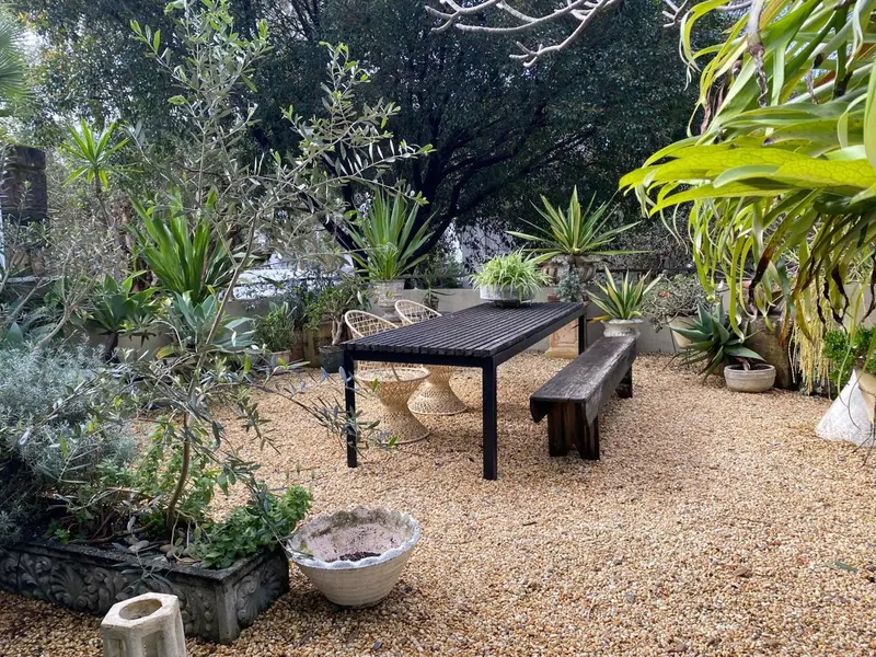 Une table et un banc en bois sont installés sur un sol de gravier au milieu d'une végétation luxuriante.