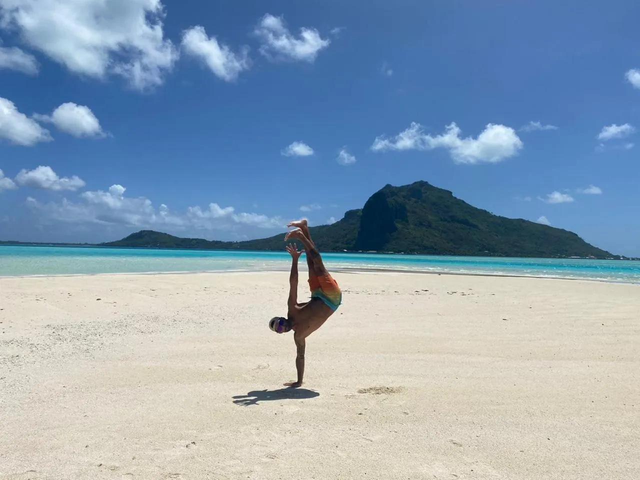 Un homme pratique une figure acrobatique sur une plage de sable blanc face à la mer turquoise.