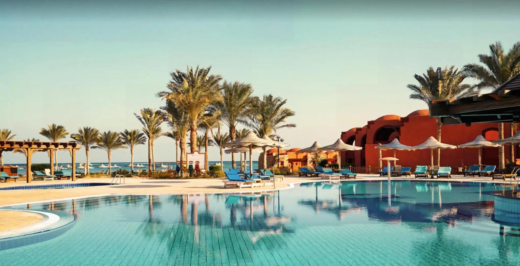 Transats bleus alignés au bord d'une piscine turquoise, avec des palmiers et des parasols en arrière-plan.