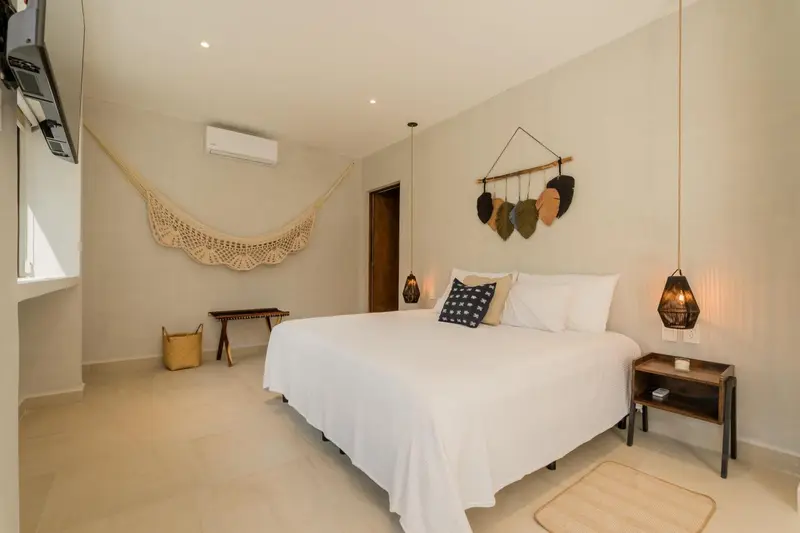 Une chambre lumineuse avec un grand lit recouvert de blanc et une décoration murale en macramé.