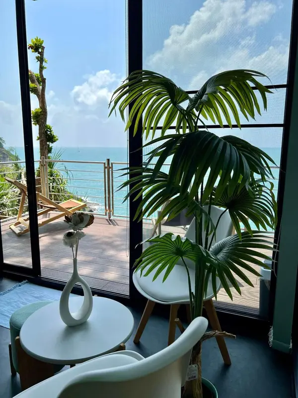 Vue sur la mer depuis un espace de détente intérieur avec mobilier clair et plantes tropicales.