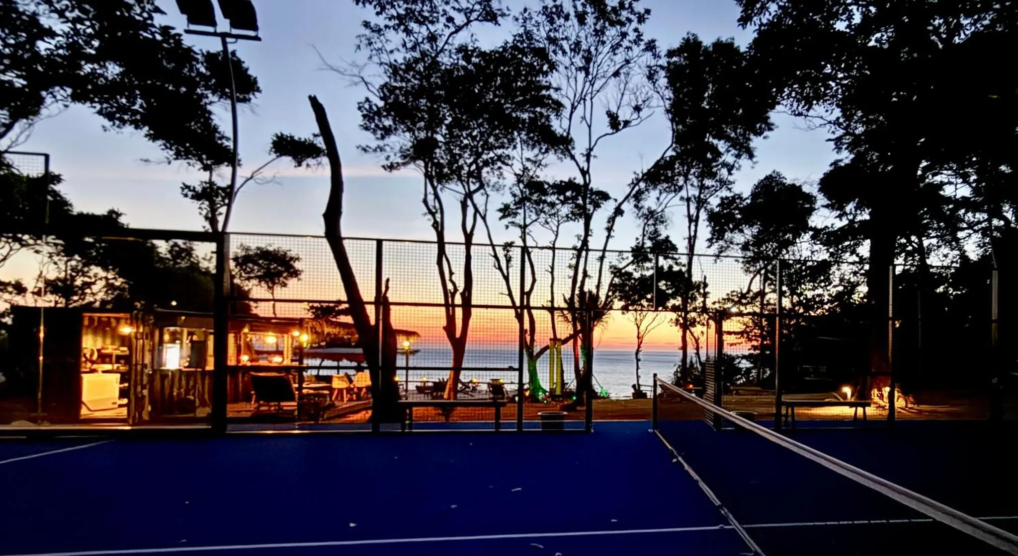 Un terrain de sport bleu au premier plan, avec un bar et des tables en arrière-plan face à la mer au coucher du soleil.