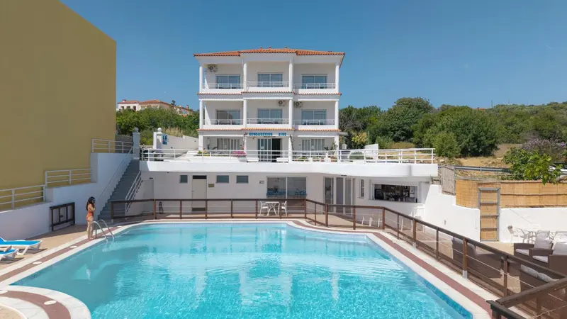 Bâtiment blanc à deux étages avec piscine au premier plan et végétation en arrière-plan.