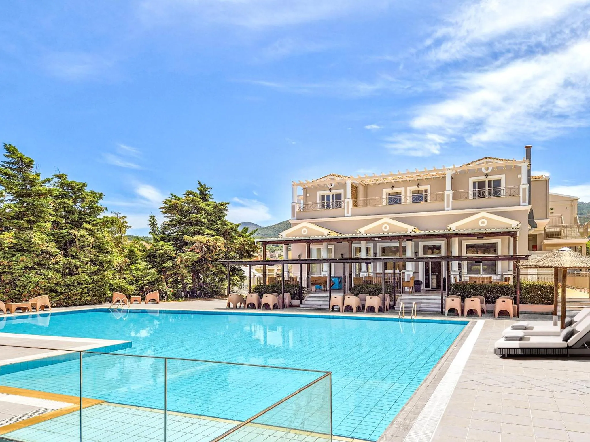 Une piscine turquoise est bordée de transats devant un bâtiment beige à deux étages.