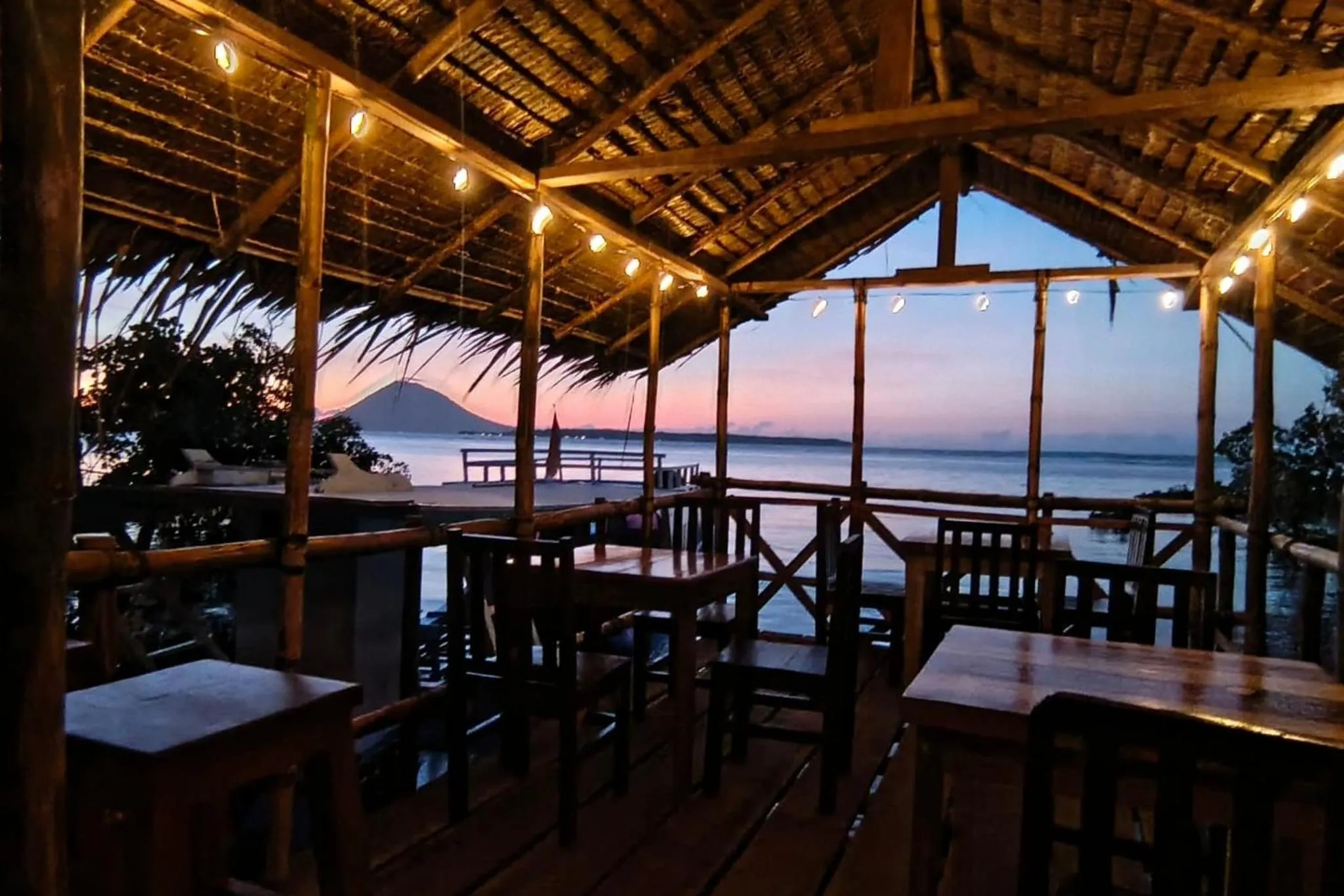 Terrasse ombragée avec tables et vue sur la mer au coucher du soleil.