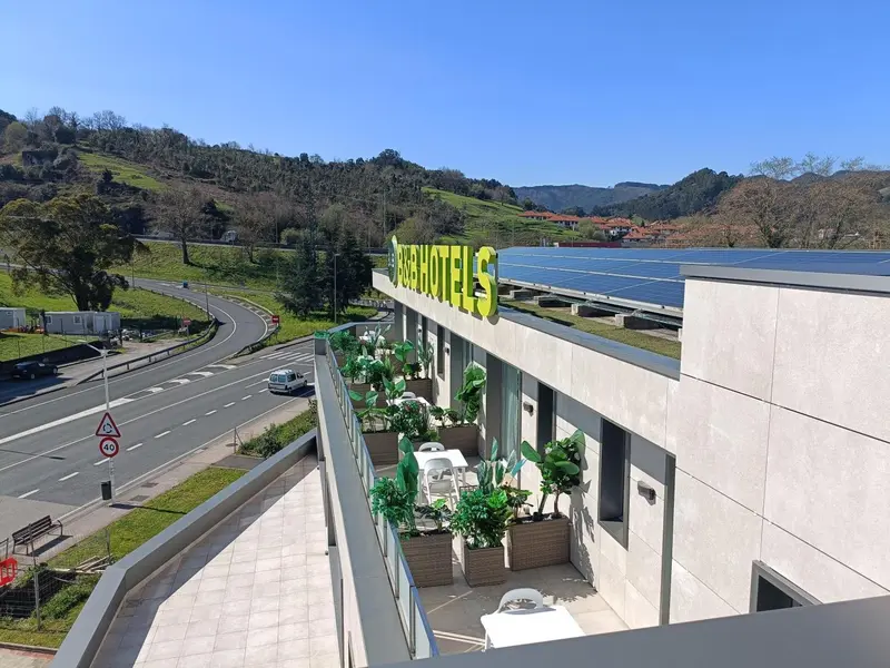 Vue d'un bâtiment moderne avec une terrasse fleurie surplombant une route et des collines verdoyantes.