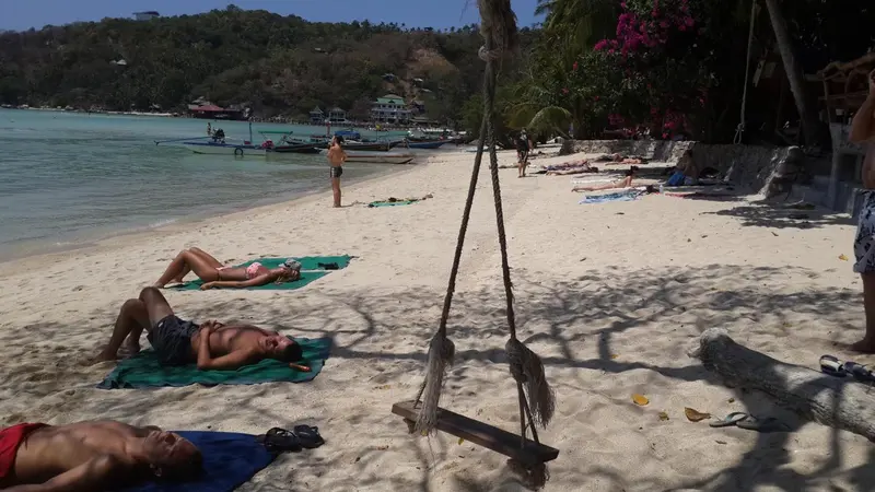 Plage de sable blanc avec des personnes se relaxant au soleil et des bateaux amarrés dans la baie.