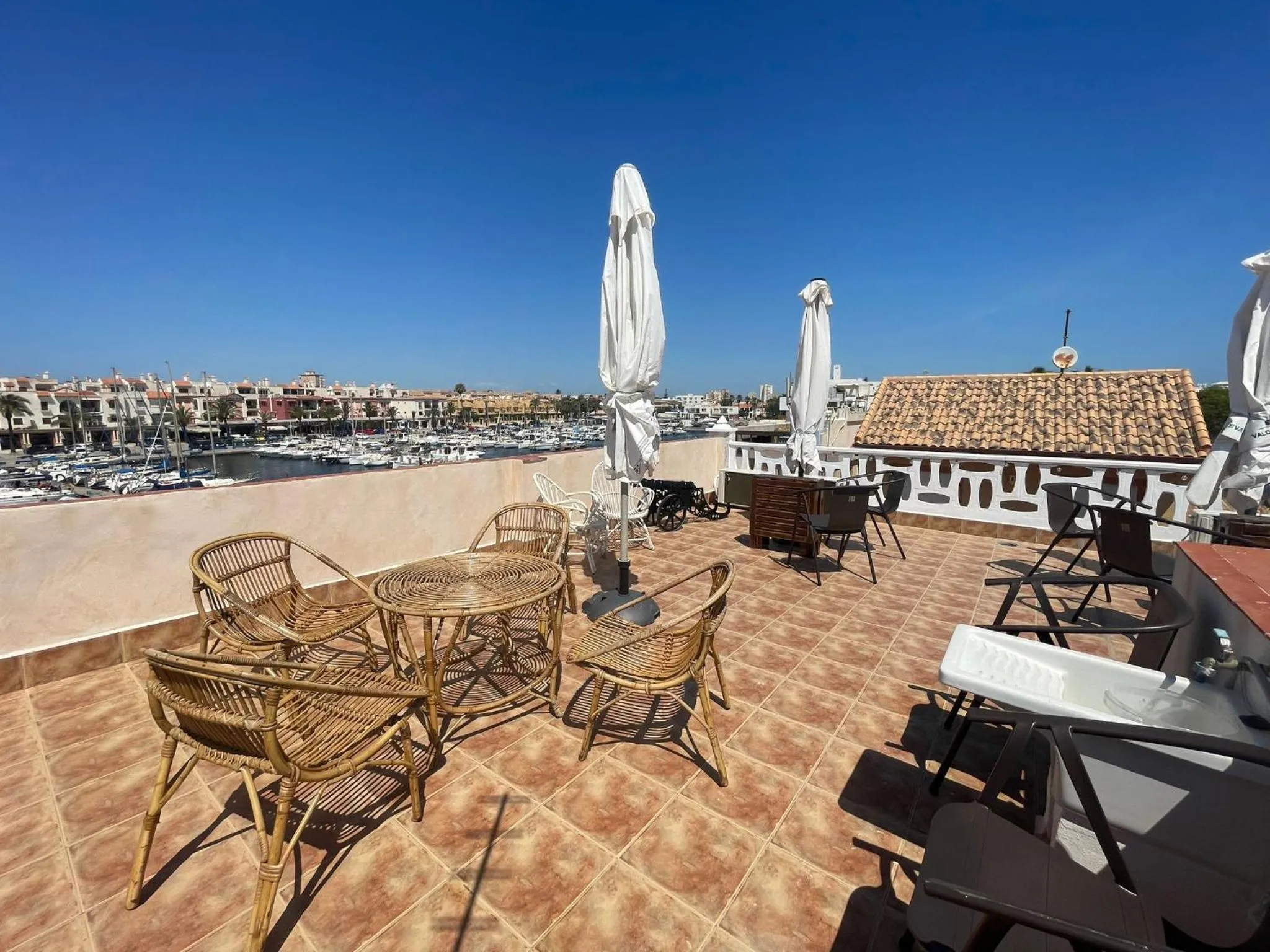 Terrasse carrelée avec mobilier en rotin et vue sur un port de plaisance ensoleillé.