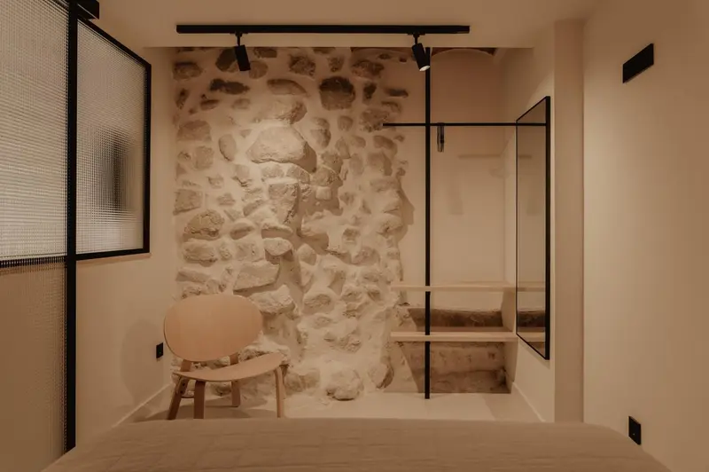 Une chaise en tissu clair est placée devant un mur de pierre brute éclairé par des spots.