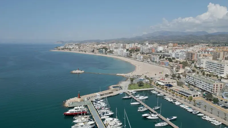 Vue aérienne d'une côte avec une plage, un port de plaisance et une ville en arrière-plan.