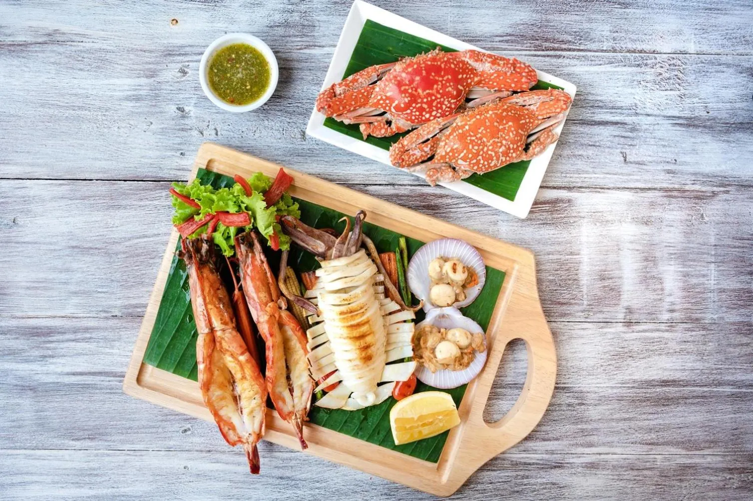 Assortiment de fruits de mer grillés présentés sur une planche en bois avec des feuilles vertes et des accompagnements.