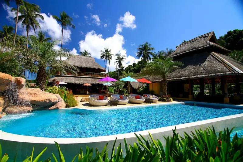 Piscine turquoise bordée de chaises longues sous des parasols colorés, dans un cadre tropical.