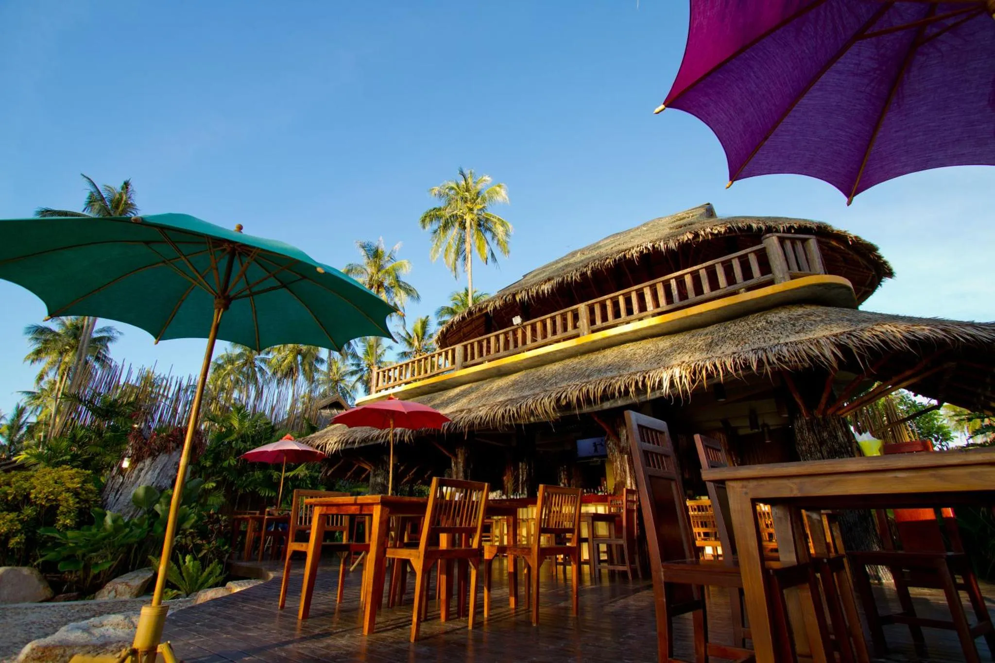 Terrasse ombragée avec tables et parasols devant un bâtiment à toit de chaume.
