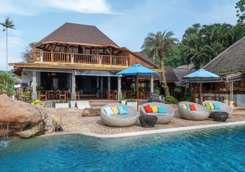 Terrasse avec mobilier d'extérieur et piscine turquoise devant un bâtiment tropical.