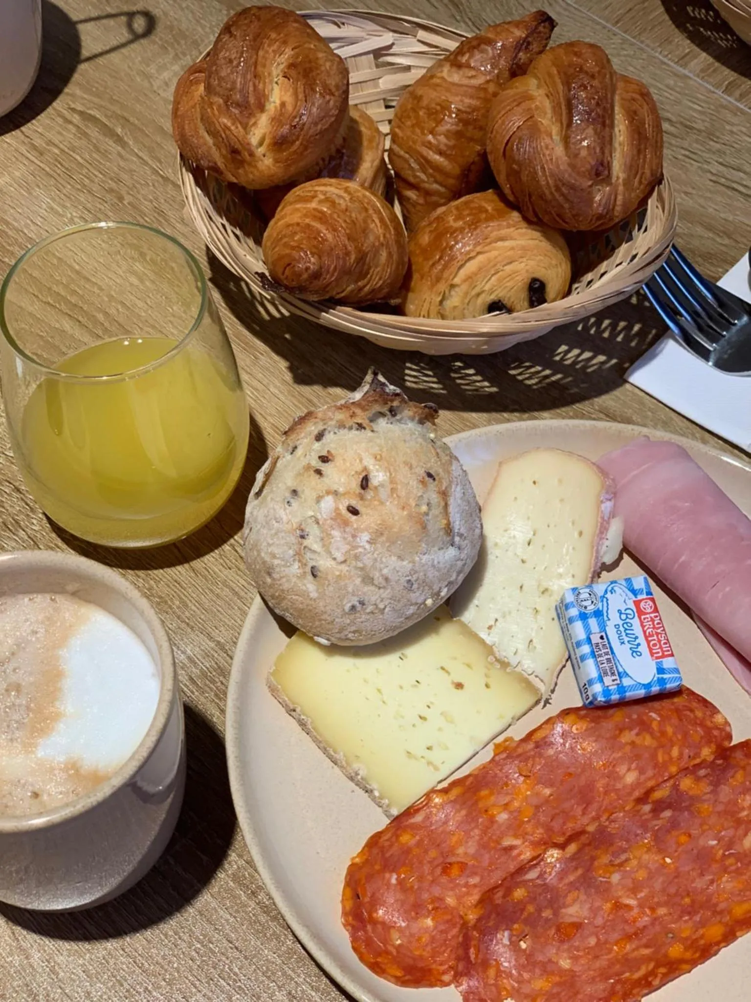 Une table présente un petit-déjeuner avec des viennoiseries, du pain, du fromage, de la charcuterie et des boissons.