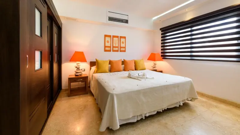 Une chambre lumineuse avec un grand lit recouvert d'une courtepointe blanche et de coussins orange.