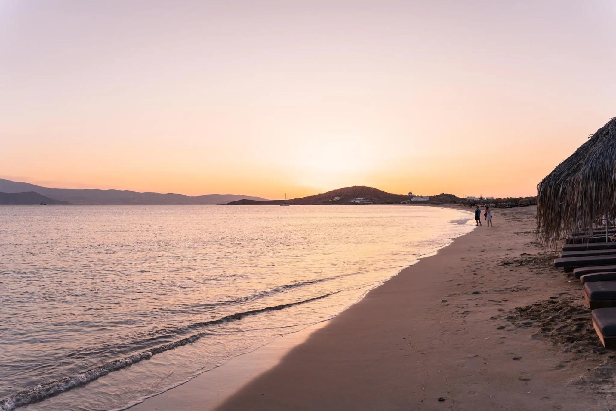 Plage de sable fin au coucher du soleil, avec des transats et une mer calme.