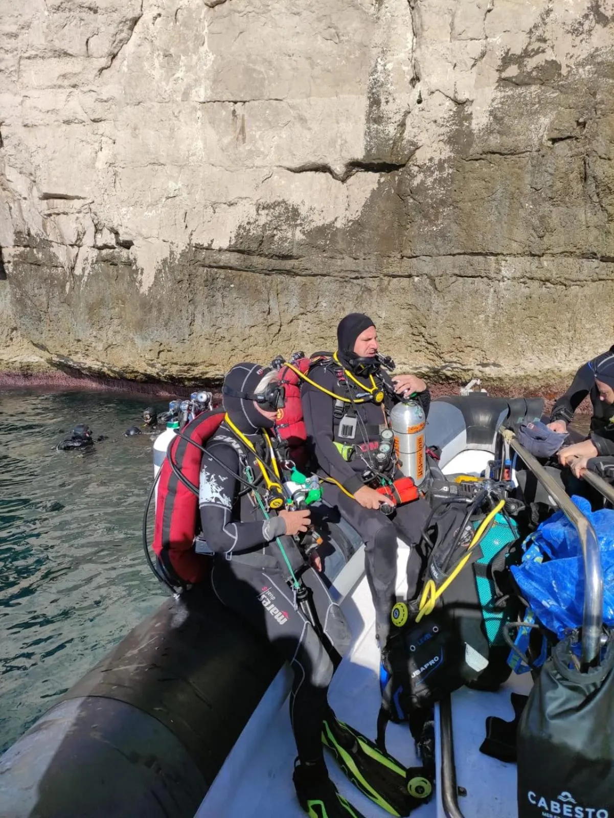 Des plongeurs équipés se préparent à bord d'un bateau pneumatique près d'une falaise rocheuse.