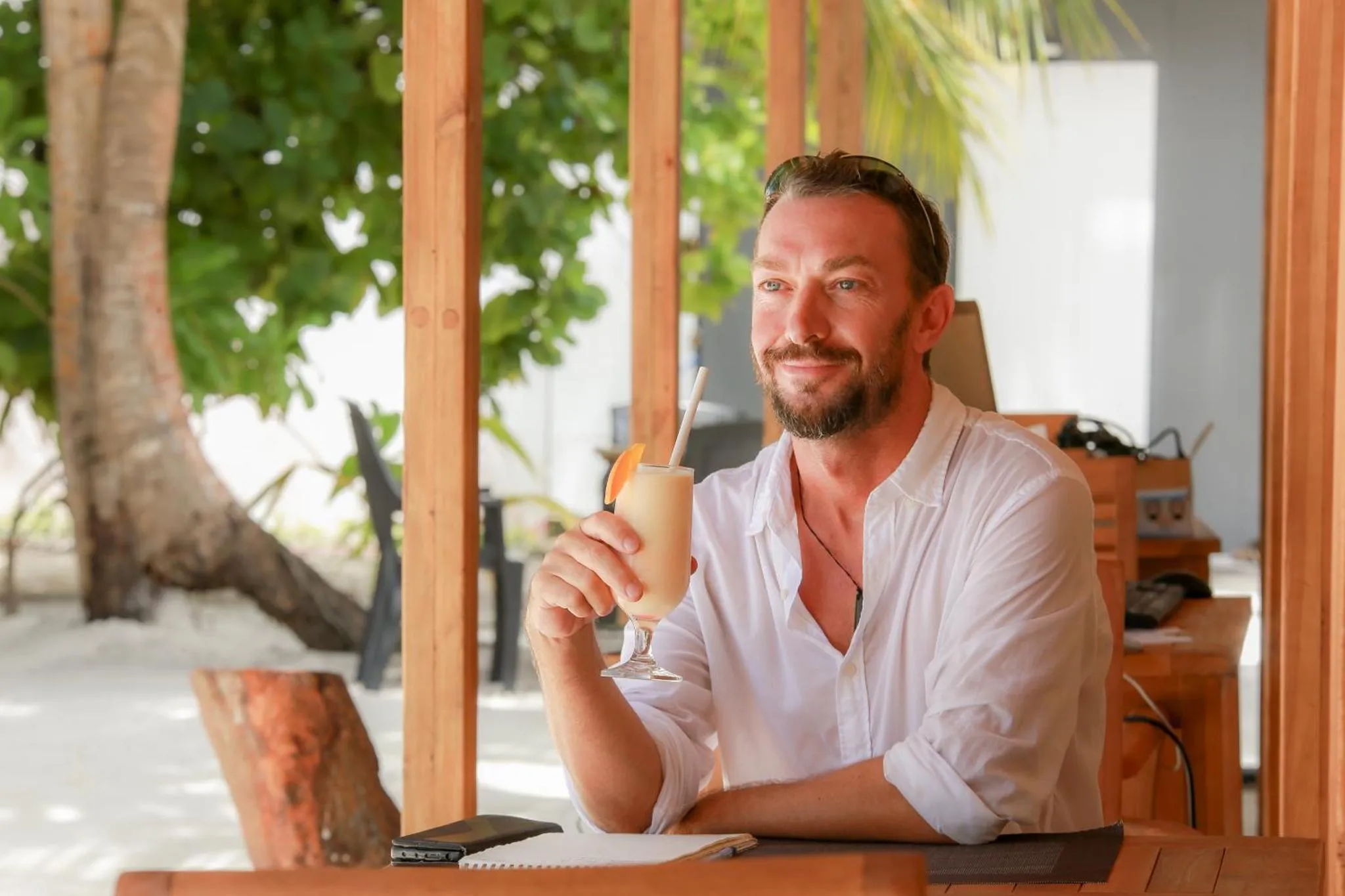 Un homme souriant tient un cocktail sur une terrasse en bois.