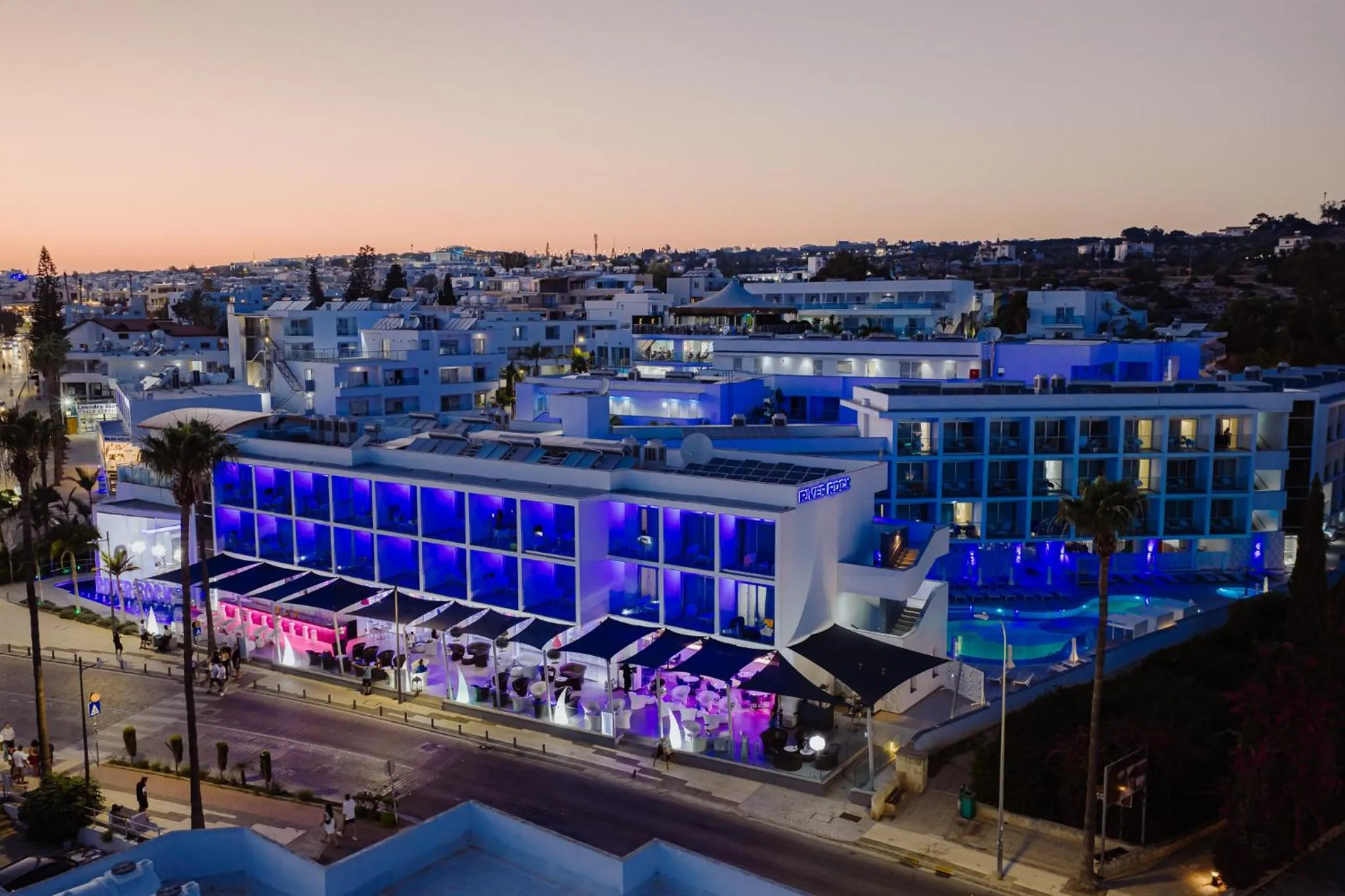 Bâtiment moderne illuminé au crépuscule avec des palmiers et une piscine.