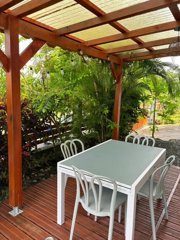 Une table et des chaises blanches sont disposées sur une terrasse en bois ombragée par une pergola.