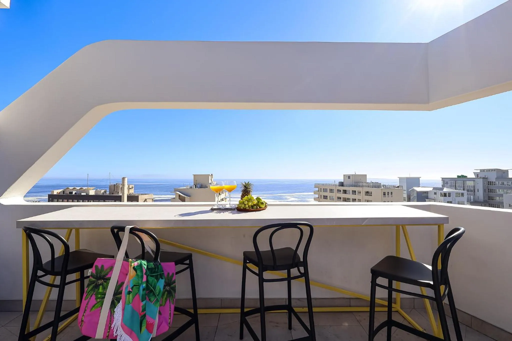 Une table avec des boissons et des fruits est mise en valeur sur une terrasse avec vue sur la mer et la ville.
