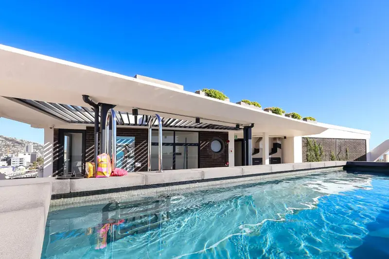 Une piscine rectangulaire avec une terrasse ombragée et une vue sur la ville.