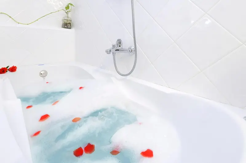 Baignoire remplie d'eau savonneuse et de pétales de roses dans une salle de bain lumineuse.