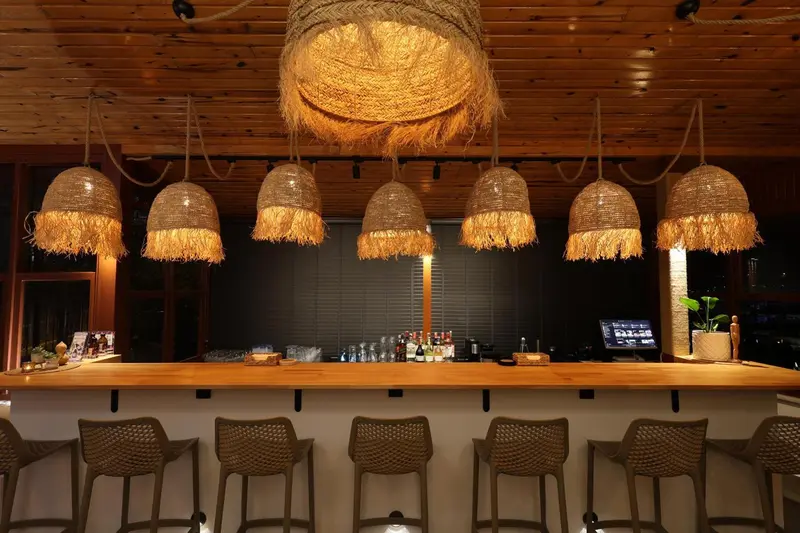 Un comptoir de bar en bois clair est éclairé par des suspensions en fibres naturelles.