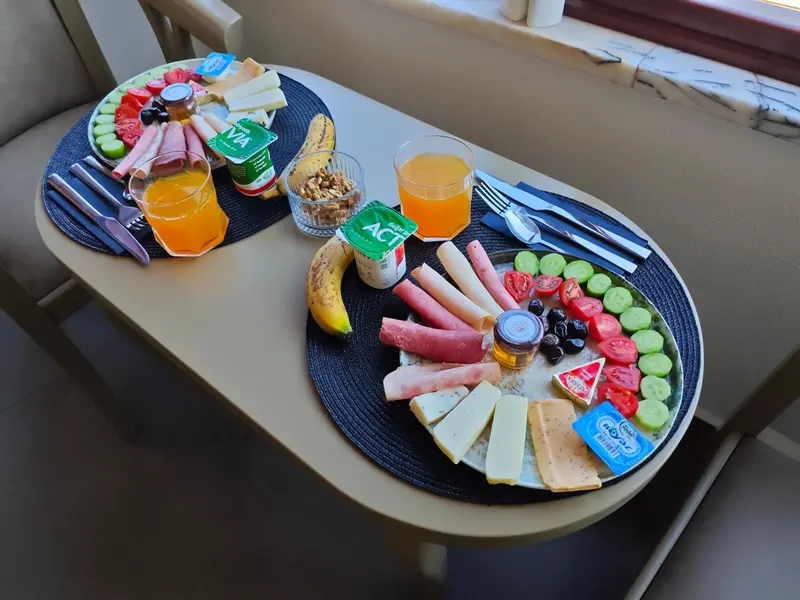 Une table est dressée avec un copieux petit-déjeuner composé de fruits, de charcuterie et de yaourts.