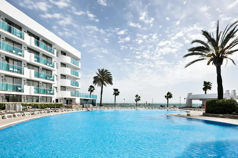 Une piscine turquoise bordée de transats devant un bâtiment blanc et des palmiers.