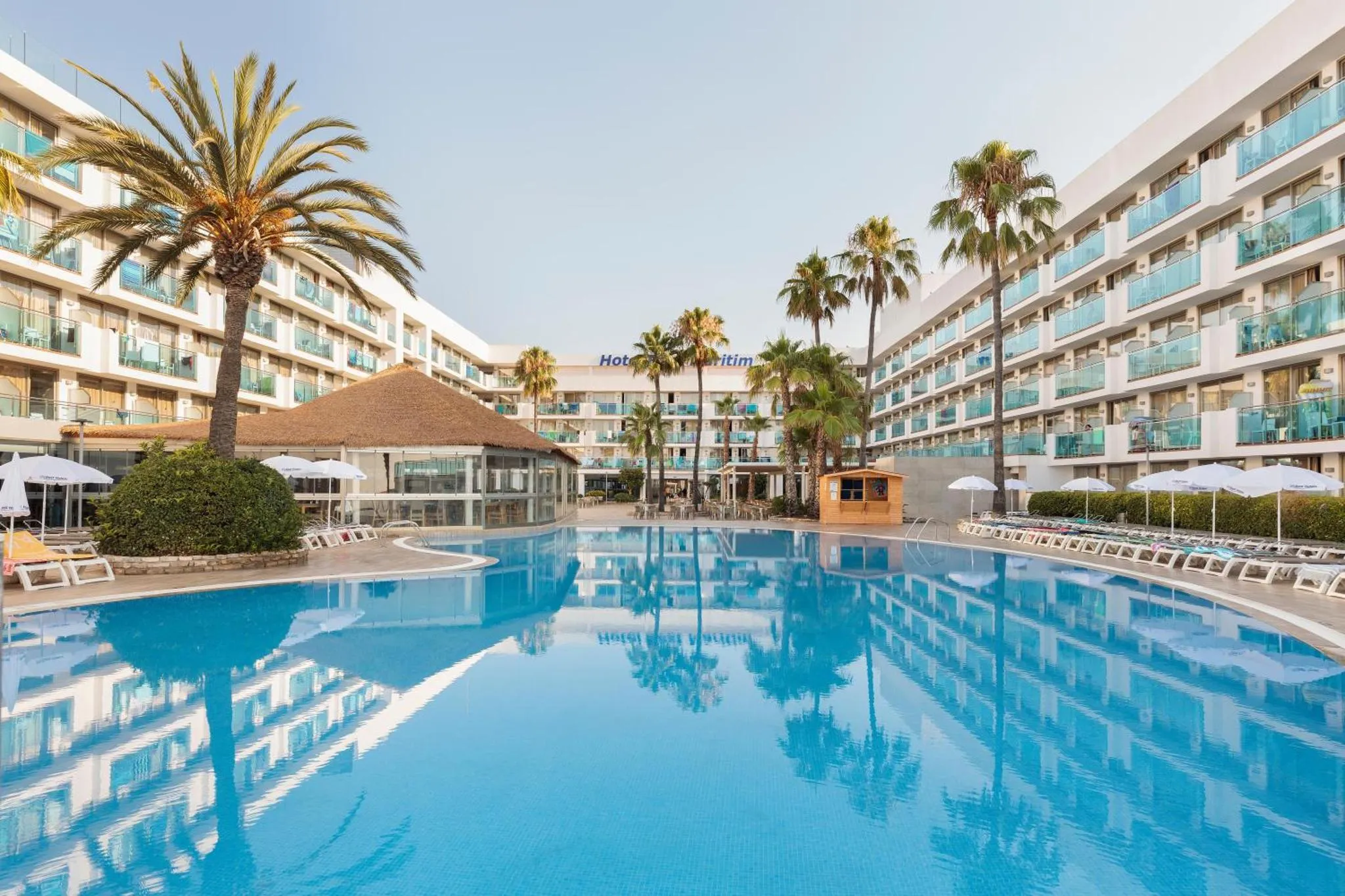 Vue d'une piscine turquoise bordée de transats et de palmiers devant un bâtiment hôtelier.