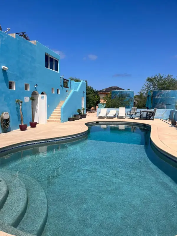 Piscine turquoise bordée d'une terrasse ensoleillée devant un bâtiment bleu.