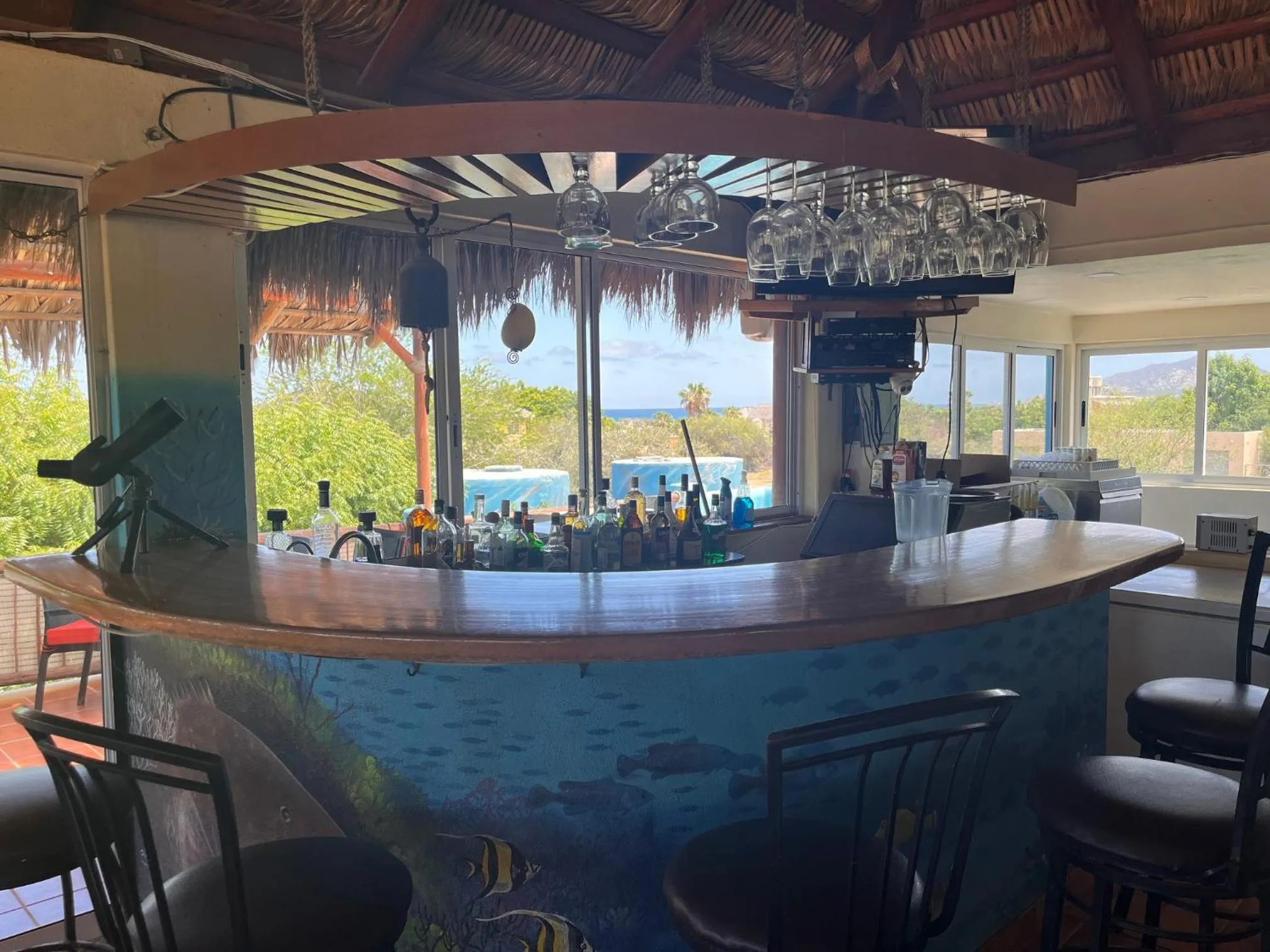 Un comptoir de bar en bois avec des tabourets, devant une vue sur la mer et une piscine.