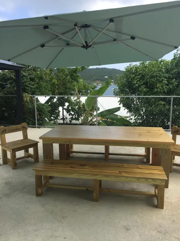 Table en bois et bancs sous un parasol sur une terrasse ensoleillée.