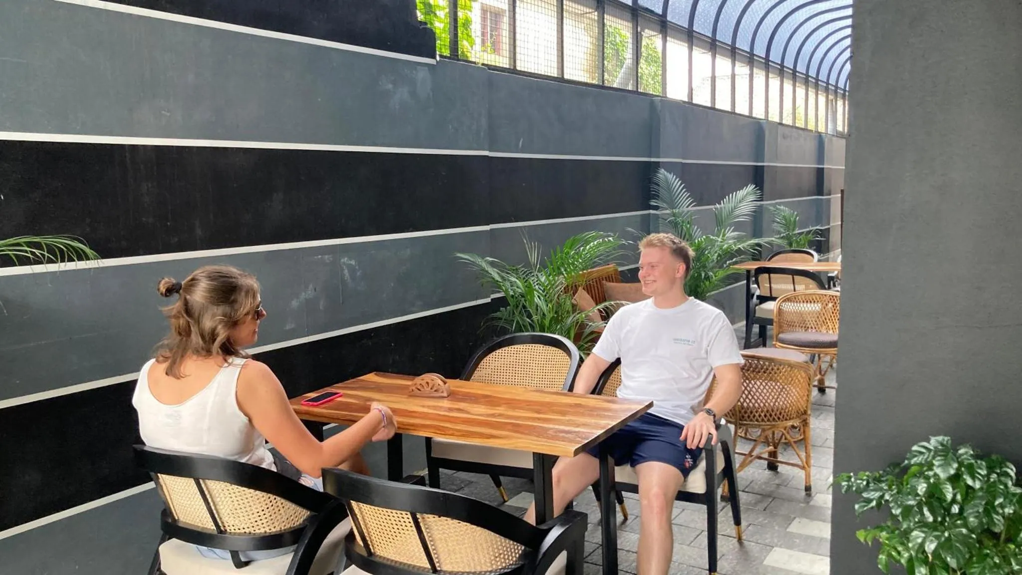 Deux personnes sont assises à une table en bois dans un espace extérieur ombragé.