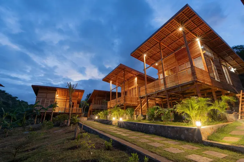 Bâtiments en bois éclairés au crépuscule avec végétation tropicale.