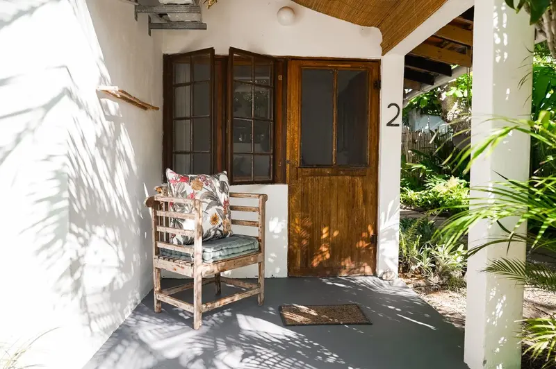 Une porte en bois s'ouvre sur une terrasse ombragée avec un fauteuil en bois.