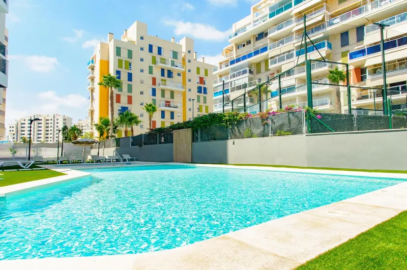 Piscine turquoise bordée d'une terrasse ensoleillée devant un bâtiment coloré.