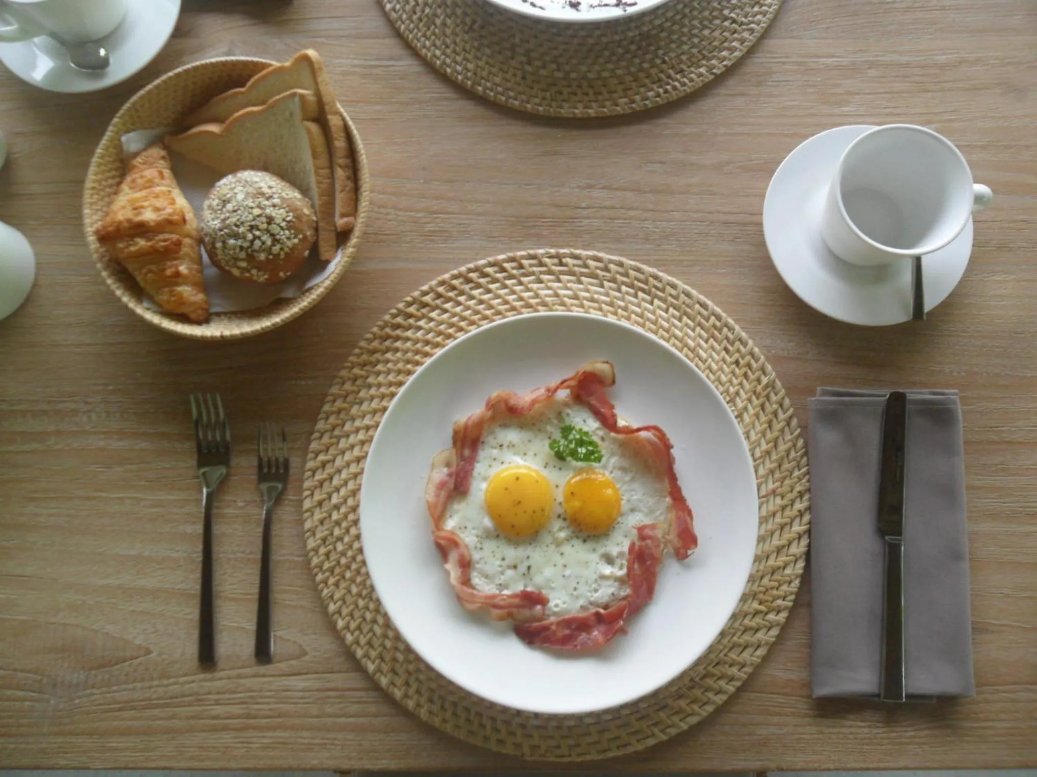Une table en bois présente un petit-déjeuner avec des œufs, du bacon et de la viennoiserie.