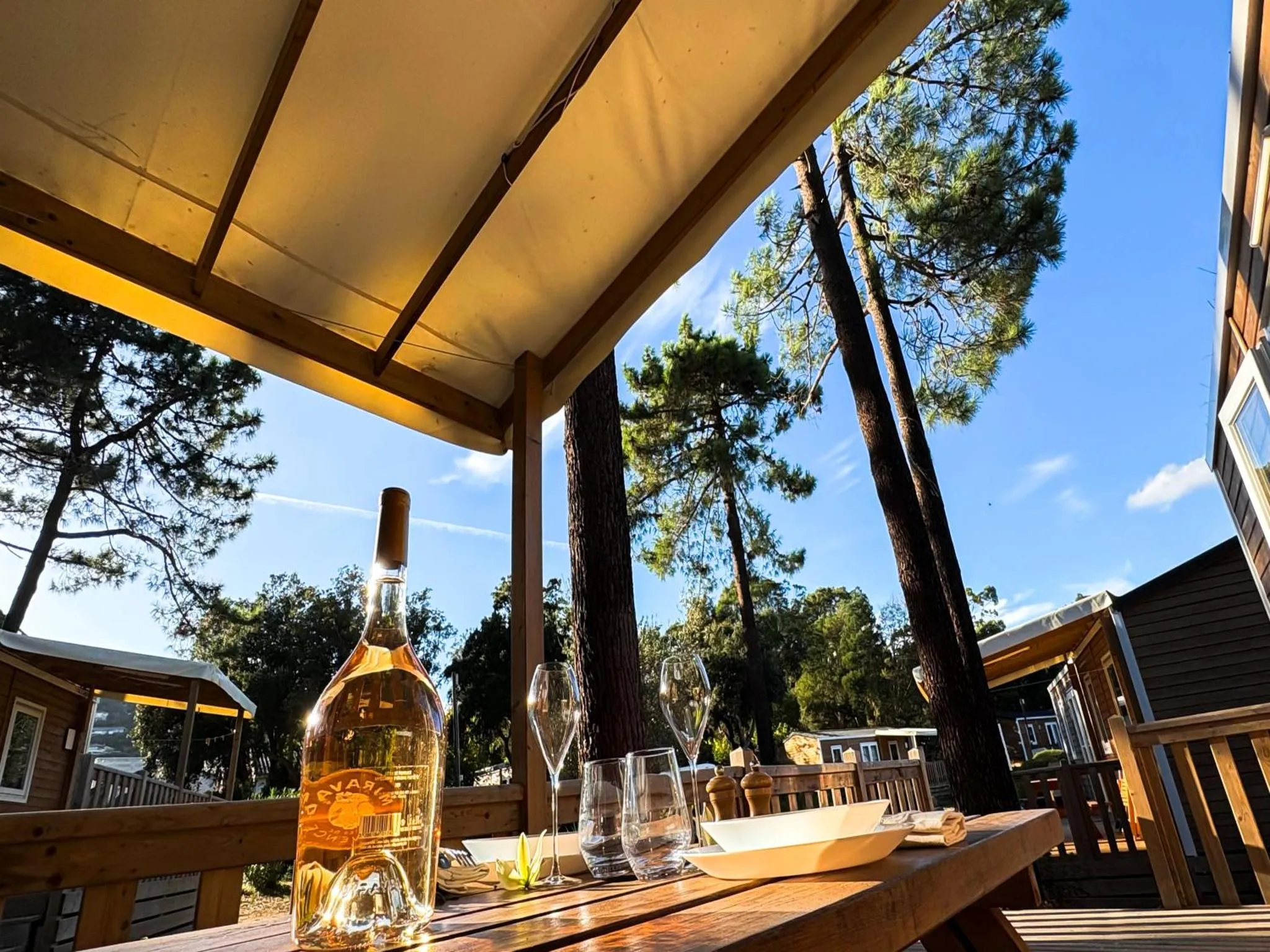 Une table en bois est dressée sous une structure ombragée avec une bouteille et des verres.